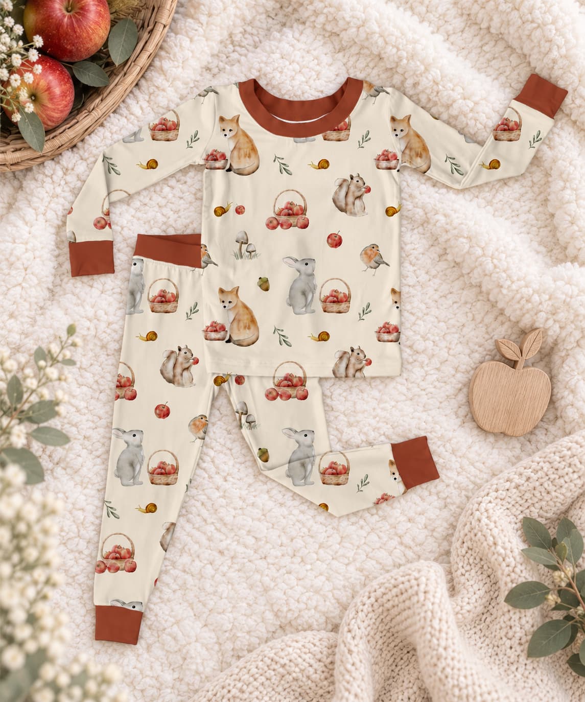 Woodland orchard bamboo pajama set for kids laid flat on a soft cream knit blanket, featuring a woodland autumn print with foxes, rabbits, baskets, apples, and leaves. The bamboo pajamas have a warm cream base, brown cuffs and collar, and are styled with seasonal props including a wooden apple, baby's breath, and a basket of apples.